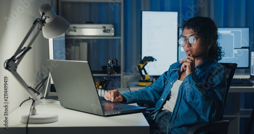 Young African American female programmer coding AI software on multiple screens in night office. Network security, cybersecurity, machine learning, working late, overtime, robotics automation concept.