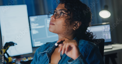 Close-up young Black female programmer coding AI software on multiple screens in night office. Network security, cybersecurity, machine learning, working late, overtime, robotics automation concept.