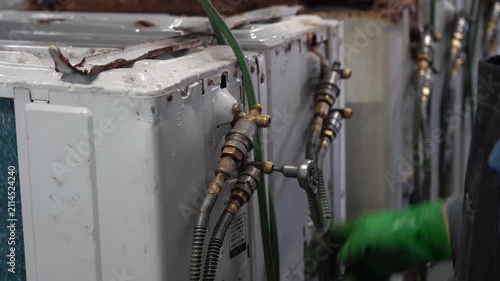 Worker in green rubber gloves connecting recovery hoses to used air conditioner units to extract refrigerant before dismantling
