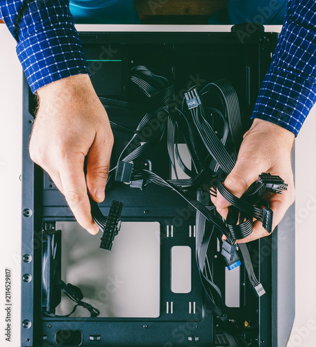 A man assembles a computer in his room at home, holding the wires from the power supply over the empty new black case lying on the table