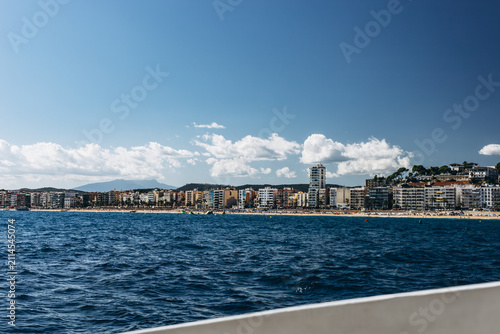 Fotografía Skyline of a coastal town spreads along the shore beyond deep blue water and white clouds