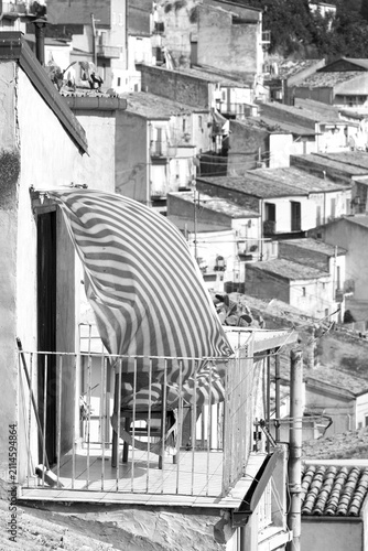 black and white small balcony with striped curtain billowing in the wind against the blurred backdrop of the houses of Leonforte, Sicily, Italy