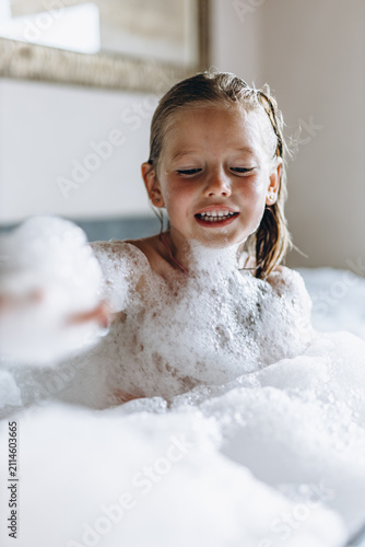 Laughing girl sits in a foamy bubble bath with a mound of soft foam on top of her head. Childhood bath time joy and gentle hygiene for kids bath care and family wellness brands