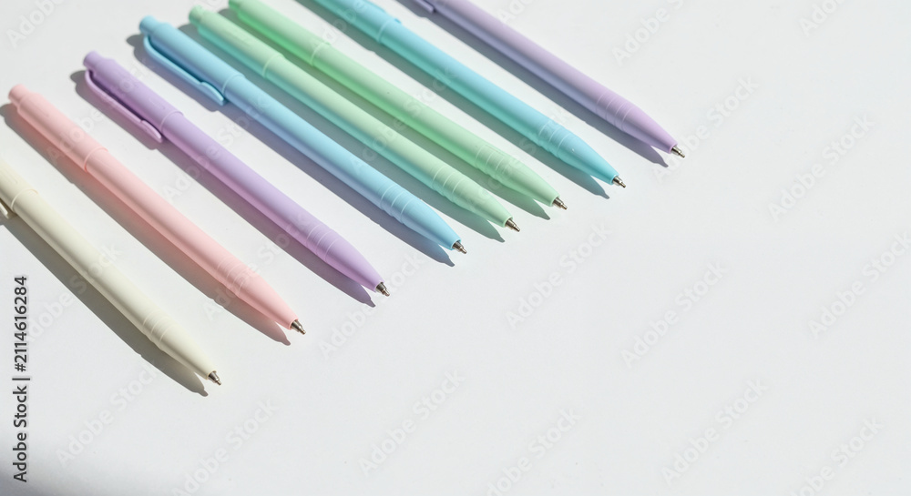Fototapeta premium High angle view of pastel-colored ballpoint pens lined up in a neat diagonal row casting soft directional shadows on a clean white background for modern office aesthetic.
