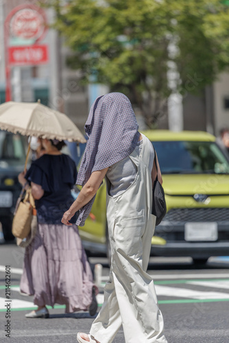Side profile of a person in white overalls covering their head with a plaid shirt for sun protection while crossing a city crosswalk during extreme summer heat.