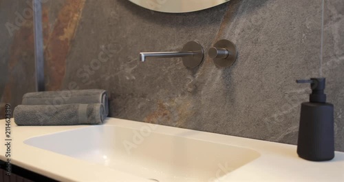 View of luxury modern bathroom with built in wall faucet and stone basin, slow camera panorama reveals dark natural stone cladding wooden vanity rounded mirror and dark accessories shaping interior