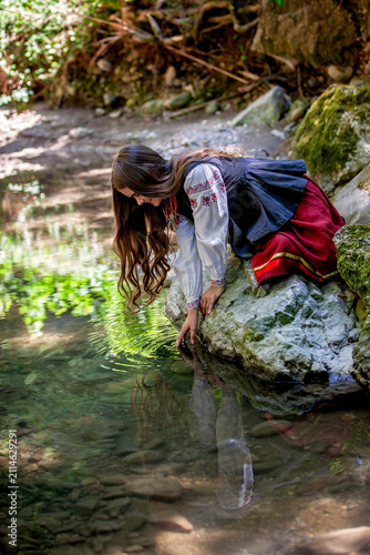 Peaceful young Ukrainian woman relaxing beside a forest stream in traditional embroidered clothing, expressing inner balance, cultural identity and harmony with nature.