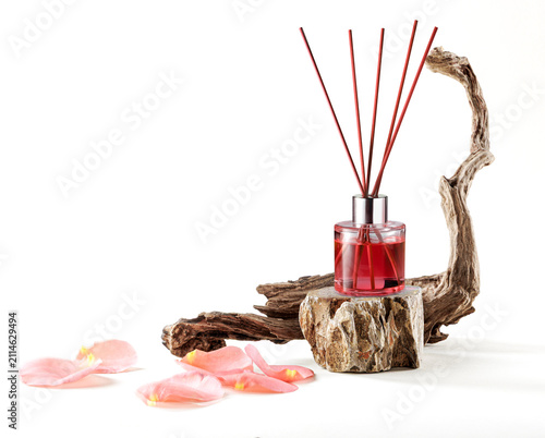 Rose essence reed diffuser bottle on a stone stand beside driftwood and rose petals isolated on blank white background.