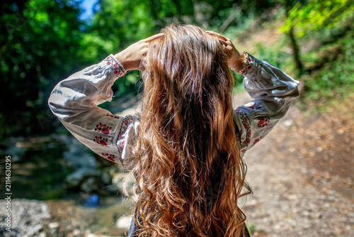 Young Ukrainian woman in embroidered clothing showing her long natural hair in a sunlit forest, combining feminine beauty, cultural tradition and nature.
