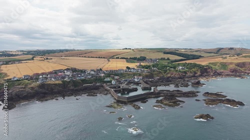 Aerial drone flight directly forward over the historic fishing village and harbour of St Abbs, revealing the scenic rugged Berwickshire coast and rural rolling fields under a cloudy sky in Scotland.