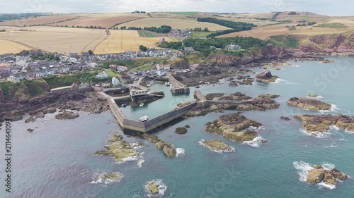Aerial view of the fishing harbour, pier, and coastal village of St Abbs, highlighting the rocky shore and scenic countryside in Berwickshire, Scotland.