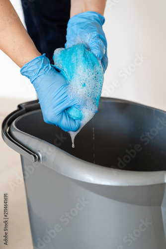 Close-up of hands in blue nitrile gloves wringing out a soapy microfiber cloth over a plastic bucket. Professional cleaning service or chores concept.