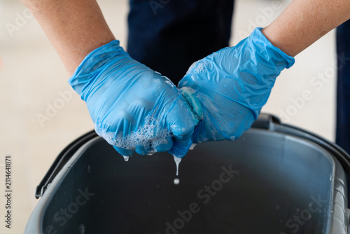 Close-up of hands in blue nitrile gloves wringing out a soapy microfiber cloth over a plastic bucket. Professional cleaning service or chores concept.