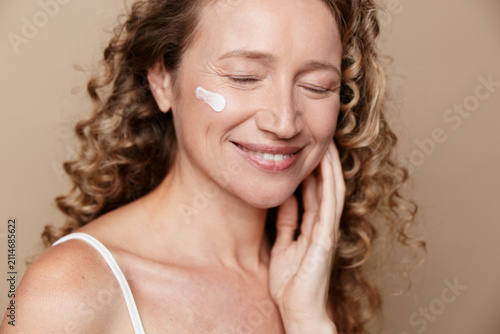 Foto Authentic middle-aged woman with natural skin smiling gently while applying cream on cheek