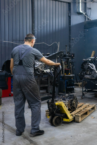 Man mechanic relocates large engine toward diagnostic section of workshop. Worker uses industrial forklift to transport engine. Vertical shot