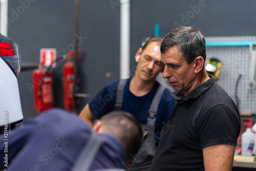 Mature mechanic with colleagues examine car damage in repair shop. Employee team fixes broken vehicle at service station. Specialists group