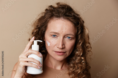 Obraz na plátne Natural looking woman applies facial moisturizer on a beige background showcasin
