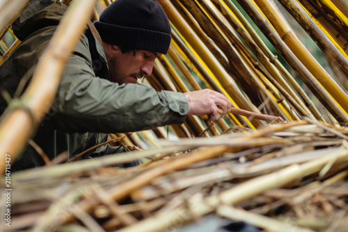 Man wearing a beanie and jacket focused on constructing a makeshift shelter from bamboo and branches
