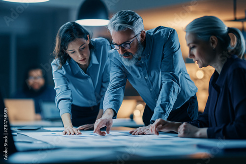 Three professional colleagues collaborate in a dimly lit office leaning over a large document on a table to analyze complex data and strategize together