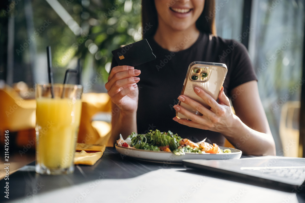 Obraz premium Young woman using smartphone and credit card to pay for healthy meal at a restaurant table with salad and orange juice in a bright, modern setting
