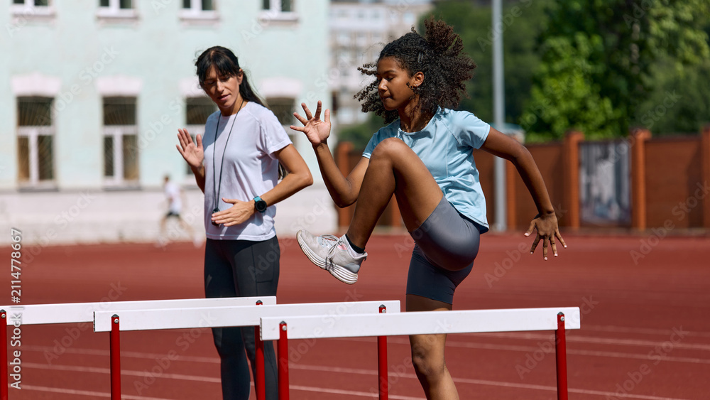 Fototapeta premium Teenage female athlete practicing hurdle approach while adult female coach instructs. Concept of hurdle preparation routine, youth athlete instruction, summer sport skill building.