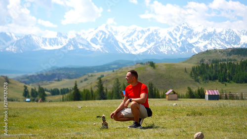 Fotografía Inspired athletic traveler kneeling on a mountain ridge and carefully building a balanced zen stone pyramid, captured from a dramatic low point of view against a majestic snow-covered ridge