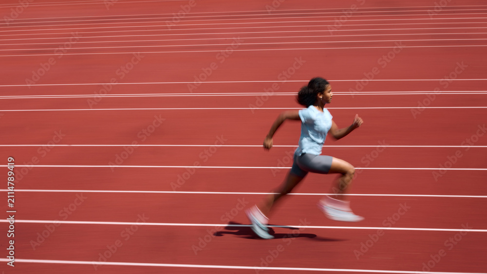 Fototapeta premium Side view of Black teenage female runner accelerating across red running track with motion blur. Concept of sprint speed development, youth athletics training, summer sport performance.