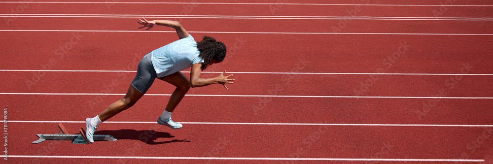 Fototapeta premium Teenage female runner launching from low crouched sprint start on track. Concept of sprint start technique, athletic readiness, summer sport performance. Banner