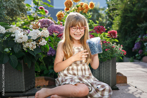 Little schoolgirl with glasses picking and eating fresh ripe blueberries in summer. Happy child enjoying nature. Real life, real people.
