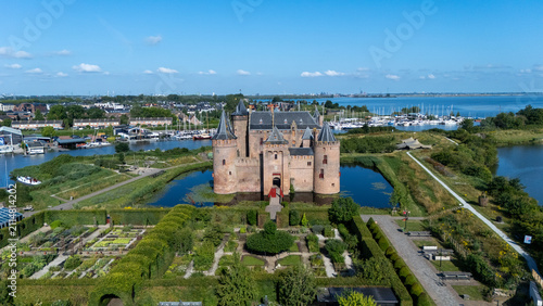 Aerial view of a historic medieval castle surrounded by water, gardens and green landscape. Scenic travel image showing fortress architecture, towers, moat, marina and blue sky near a Dutch waterfront