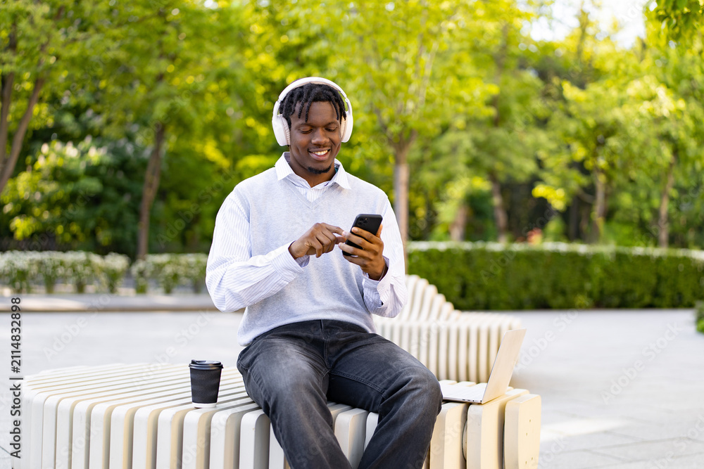 Obraz premium A smiling young African American man wearing headphones sits on a modern bench in a park, looking at his phone with a laptop nearby