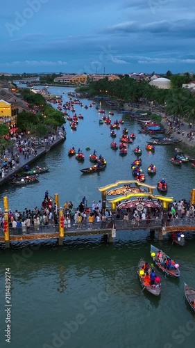 Vertical aerial drone video of Hoi An Ancient Town at dusk, Vietnam, featuring colorful lantern boats, crowded riverside promenade, and vibrant tourism atmosphere along the Thu Bon River.