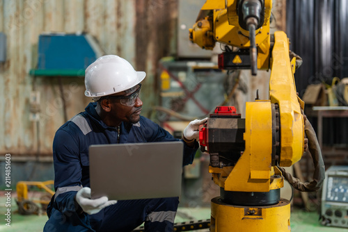 Worker wears a white hard hat and dark blue work shirt. He holds a laptop near a yellow robotic arm. Worker checks the arm's components with white gloves. Industrial background features metal walls