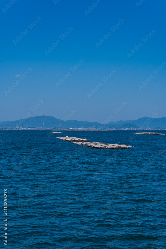 Naklejka premium Floating oyster rafts on calm blue sea with distant mountains under clear sky, showcasing coastal aquaculture, marine landscape, and peaceful waterfront scenery