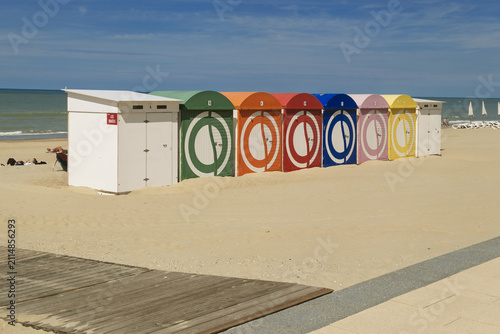 Foto A row of colorful beach huts stands on the sand of Malo-les-Bains beach in Dunkirk, their hues contrasting with the clear sky and the low tide