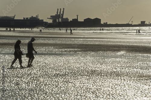 Fotografía Silhouettes of two people walk on the beach at Dunkirk at low tide, the wet sand reflecting the light while the distant port appears on the horizon