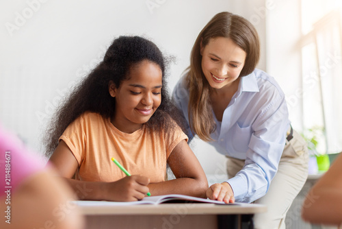 Bild auf Leinwand Supportive female teacher helping black schoolgirl complete written classwork at