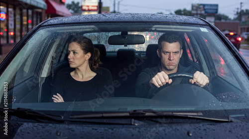 Man and woman sitting in a car during rain, expressions of tension visible, city street and storefronts blurred in the background