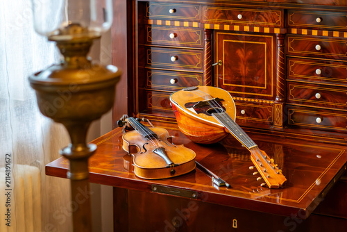 Quadro su tela Vintage violin with bow and mandolin resting on an open inlaid wooden secretary desk with small drawers, with a brass oil lamp in the foreground of an elegant retro interior