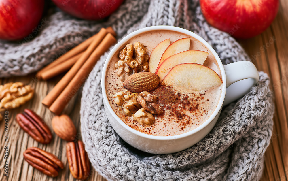 Fototapeta premium Top view of hot chocolate drink in white mug topped with fresh apple slices walnuts, cinnamon powder. Cozy winter beverage sits on wooden table next to red fruit, knitted scarf.