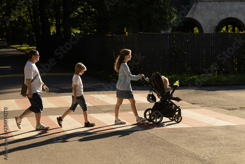 Four people, a man, a woman, a boy, and a child in a stroller, cross the road at a pedestrian crossing at sunset. Family walks around the city, safe crossing