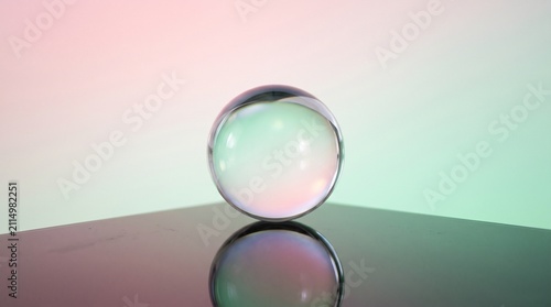 a clear transparent glass sphere resting on a reflective surface against a smooth pastel gradient background in soft pink and light green tones, creating a minimalist and serene concept.