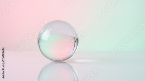 a clear transparent glass sphere resting on a reflective surface against a smooth pastel gradient background in soft pink and light green tones, creating a minimalist and serene concept.