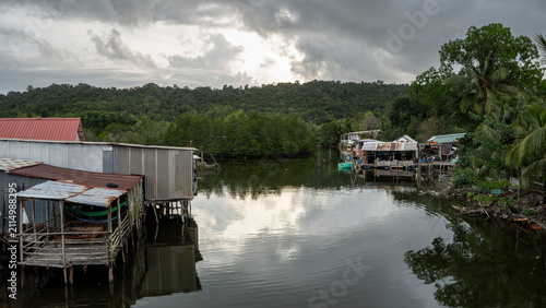 Fotografie A quiet Vietnamese fishing village with weathered stilt houses lining a calm waterway, flanked by mangroves and dense jungle hills under a dramatic overcast sky