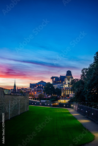 Foto Dusk view along the outer walls of the Tower of London, with a green moat lawn, Tower Bridge and city buildings glowing warmly in the background under a vivid pink-and-blue sky