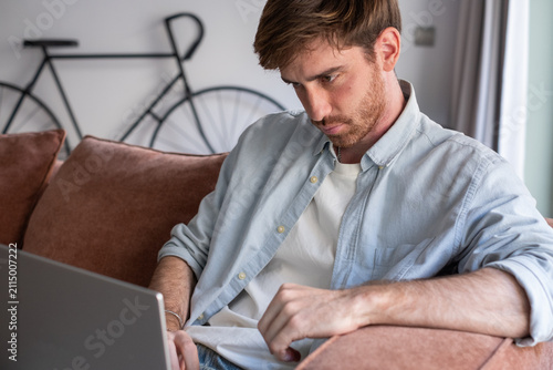 Картината върху платно Young adult man working from home, sitting on a comfortable couch and looking at