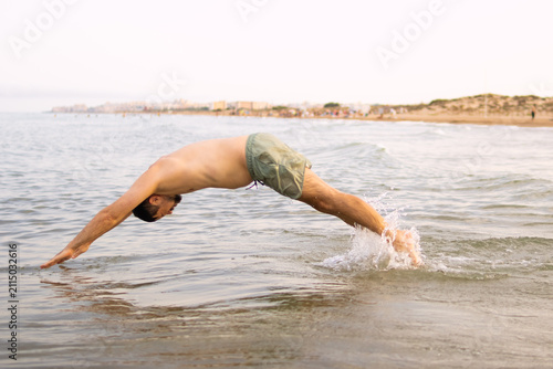 Young man diving headfirst into calm ocean water, creating a splash with his feet, enjoying summer vacation on a sunny beach day with distant coastline buildings