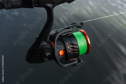 Fotografía Fishing reel closeup with green lime line, spinning rod reel mechanism, angler sportfishing gear and tackle equipment over lake water