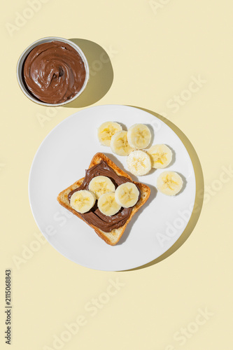 Toasted bread with chocolate cream and banana on a white plate on a yellow background