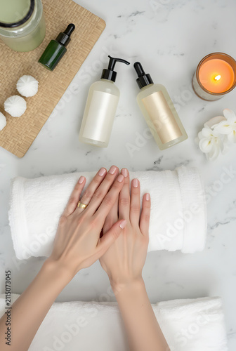 Top-down view of well-manicured hands resting on rolled white towels, surrounded by luxurious skincare bottles, dropper serum, lit candle, and flowers on a marble surface